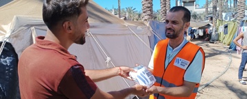 giving sadaqah last 10 days of ramadan humanitarian worker in an orange vest distributes food to a man in a refugee camp with tents and palm trees in the background