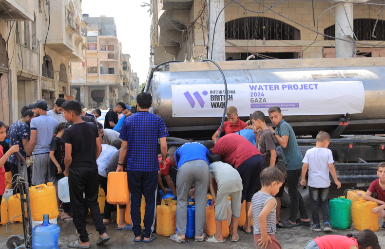 what is the meaning of waqf A group of people, including children and young men, gather around a water tanker in a damaged area, filling plastic containers with water. The banner on the tanker indicates a 2024 water project in Gaza, funded by "International British Waqf".
