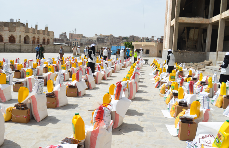 Rows of food aid packages, including sacks of grains, cooking oil, and dates, arranged for distribution in an urban area with volunteers in uniforms assisting.