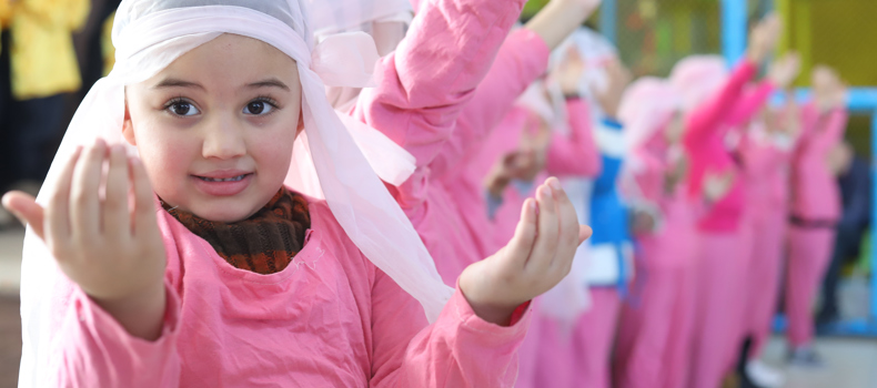 Best charity for orphans A young girl dressed in pink with a white headscarf performs a group dance or activity, with other children in matching outfits raising their hands in the background.