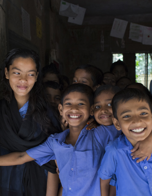 Best charity for orphans A group of smiling school children standing in a doorway, with one older girl standing beside them, all wearing blue uniforms in a classroom setting.