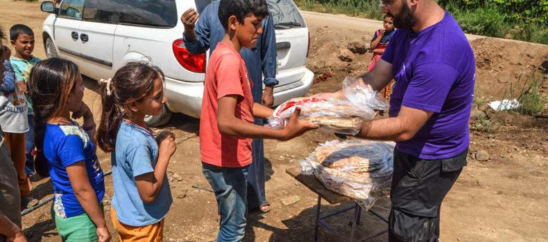 A humanitarian worker in a purple shirt distributing bread to children in a rural setting, with a white van and greenery in the background.