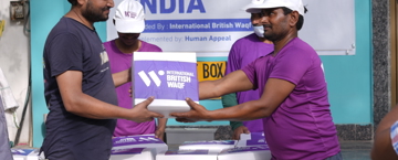 Difference Between Waqf And Sadaqah A man in a purple shirt and white cap hands a purple box labeled "International British Waqf" to another man in a navy shirt during a Qurbani 2024 distribution event in India. A banner and more distribution boxes are visible in the background.