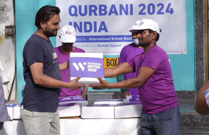 Difference Between Waqf And Sadaqah A man in a purple shirt and white cap hands a purple box labeled "International British Waqf" to another man in a navy shirt during a Qurbani 2024 distribution event in India. A banner and more distribution boxes are visible in the background.