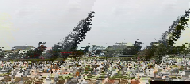 Sadaqah jariyah for parents a wide view of a Muslim cemetery with neatly arranged graves, most of them white with decorative headstones. In the background, there are trees, buildings, and a hill with a prominent white structure on top.