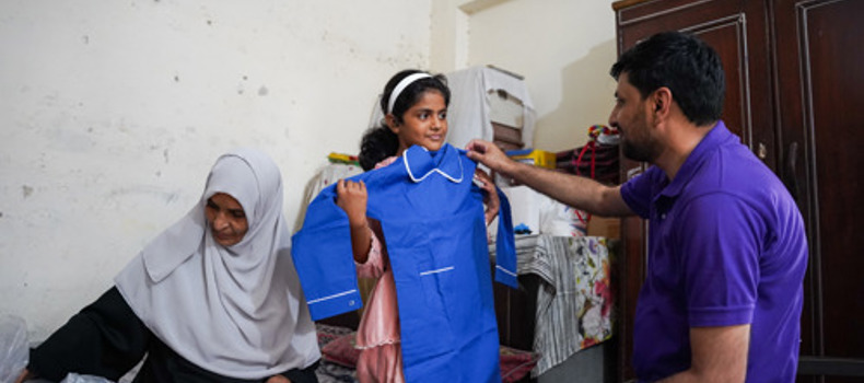 Difference Between Waqf And Sadaqah A young girl smiles while holding up a new blue outfit in a modest room, as a man in a purple shirt shows it to her. A woman wearing a light gray hijab sits nearby, writing in a notebook.