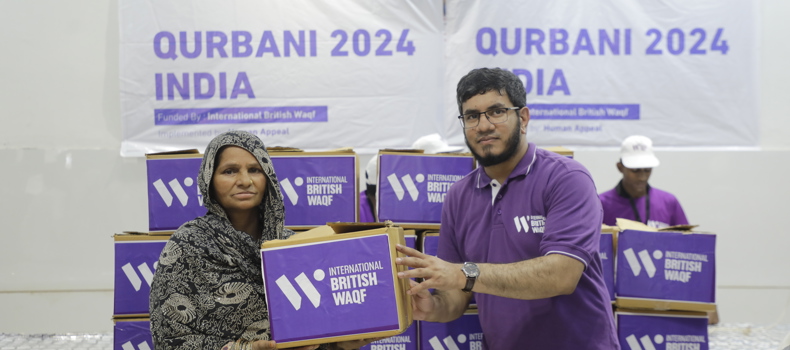 Best Form Of Sadaqah Jariyah A man and a woman hold a purple donation box labeled "International British Waqf" during a Qurbani 2024 distribution event in India. Stacks of similar boxes and a banner with the same branding are visible in the background.