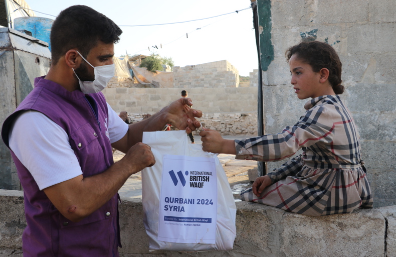 Hadith about benefits of sadaqah A humanitarian worker wearing a mask and a purple vest hands a Qurbani 2024 Syria aid bag from International British Waqf to a young girl sitting on a stone wall in a rural area.
