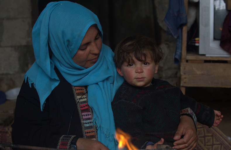 Is Giving Money To Parents Sadaqah A woman wearing a blue headscarf and a black garment with colorful embroidery is sitting beside a child who is wearing a dark sweater. There is a small fire burning in front of them. In the background, there is a wooden shelf with various items, including a television and some clothes.
