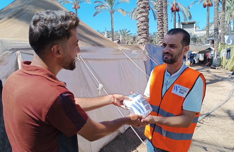 giving sadaqah last 10 days of ramadan humanitarian worker in an orange vest distributes food to a man in a refugee camp with tents and palm trees in the background