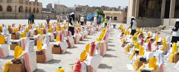 Rows of food aid packages, including sacks of grains, cooking oil, and dates, arranged for distribution in an urban area with volunteers in uniforms assisting.