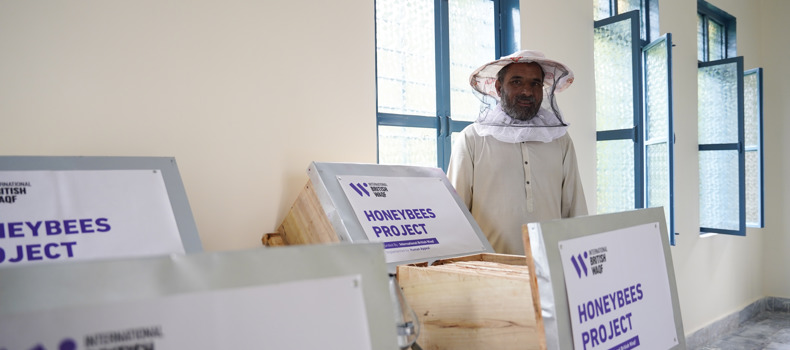 Best Form Of Sadaqah Jariyah, A person wearing a beekeeping suit stands in a room with several beekeeping boxes labeled "HONEYBEES PROJECT" by the International British Waqf. The room has multiple windows with blue frames and open shutters, allowing natural light to enter.