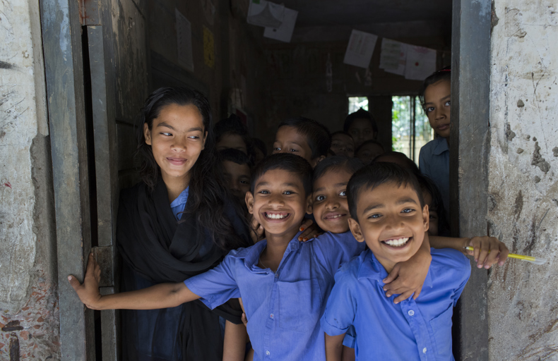 Best charity for orphans A group of smiling school children standing in a doorway, with one older girl standing beside them, all wearing blue uniforms in a classroom setting.