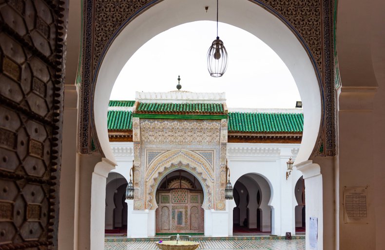 Difference Between Waqf And Sadaqah An architectural view of a mosque courtyard taken through a beautifully decorated archway. The archway has intricate patterns and designs, framing the main entrance of the mosque. The mosque features a large arch with detailed tile work, geometric patterns, and a green-tiled roof. At the center of the courtyard, there is a small fountain surrounded by patterned tiles. Hanging lanterns add to the traditional and historic atmosphere.