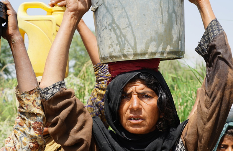 Is Giving Money To Parents Sadaqah An elderly woman carries a large metal container of water on her head, supported by her hands, while others beside her hold yellow plastic water jugs. The background shows green vegetation under a clear sky, reflecting a rural setting and the struggle for access to clean water.
