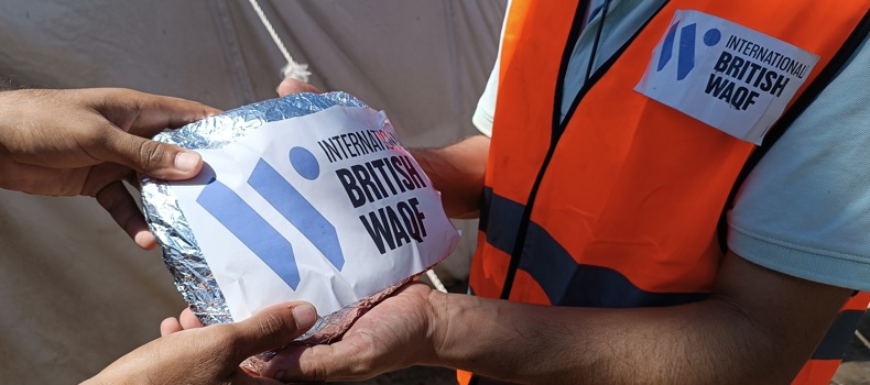 what is the meaning of waqf A humanitarian aid worker wearing an orange vest with an "International British Waqf" badge distributes a hot meal wrapped in foil to a person in need. The meal package also has the "International British Waqf" logo on it. The background features a tent, indicating an aid distribution setting.