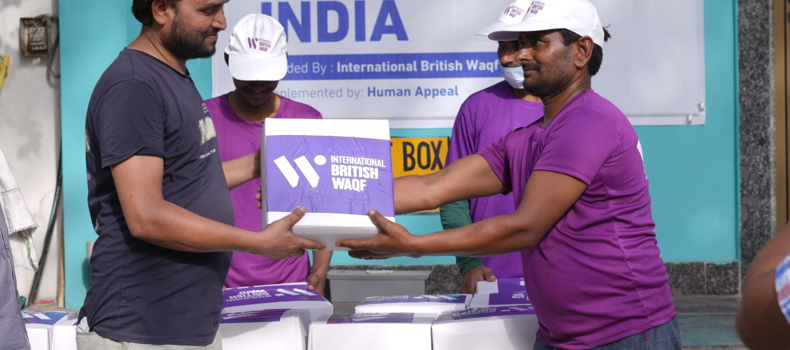 Hadith about benefits of sadaqah A volunteer in a purple shirt and white cap hands over a Qurbani 2024 food box from International British Waqf to a man during a distribution event in India. A banner and more boxes are visible in the background.
