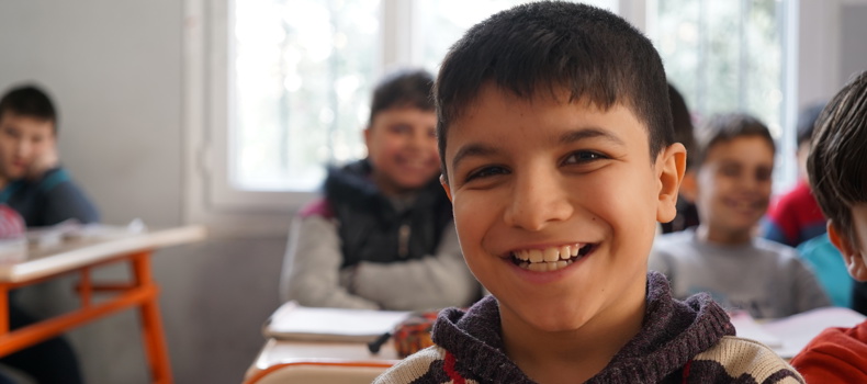 A smiling boy in a classroom, with other students sitting at desks in the background.