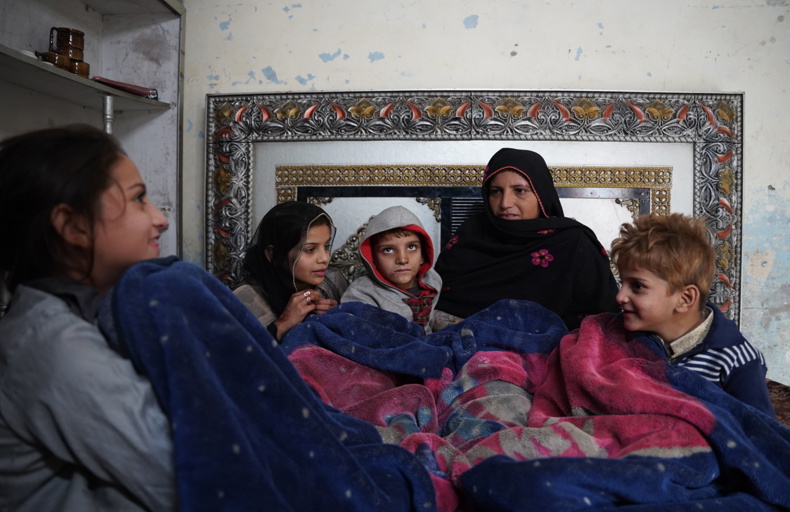 sadaqah jariyah for parents A group of children holding white bags with the 'Humanitarian Aid' logo, standing in front of a banner that reads 'British International Waqf.' In the background, there's a mural depicting two children in school uniforms and a tree, suggesting the setting might be a classroom or school environment. The image reflects a charitable event where children are receiving aid or gifts.